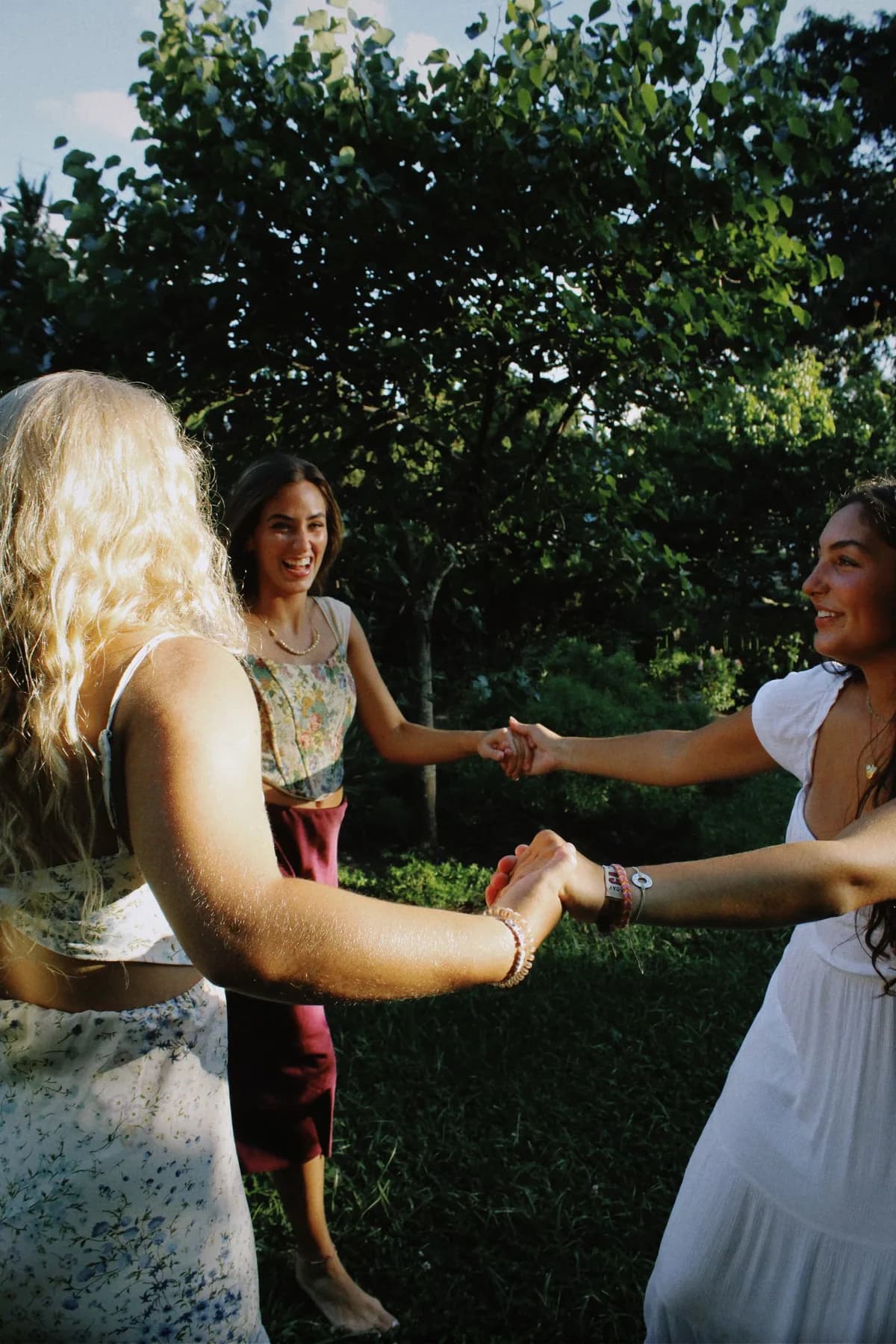 Three young women holding hands in a circle in a sunny garden, smiling at each other